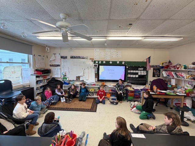 Whole-class meeting: Elementary students sit in a large circle on the classroom floor while a teacher facilitates discussion.