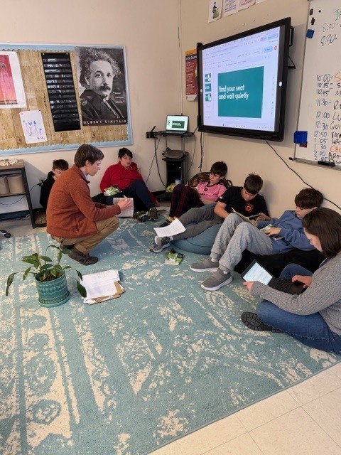Small group reading discussion: Students and a teacher sit in a circle on a rug reading and discussing books together.