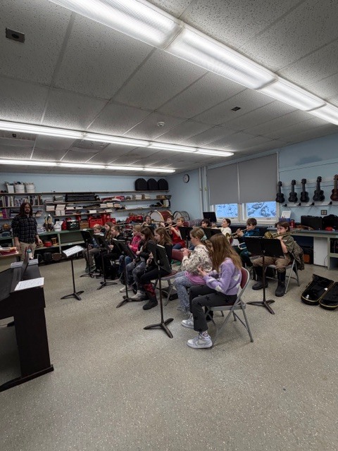 Music classroom rehearsal: Students sit on stools in a music room holding instruments while a teacher stands at the front.