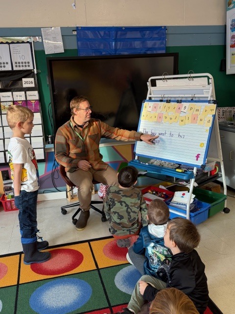 Small-group instruction on rug: A teacher sits on the floor pointing to a chart while several young students gather around and listen.