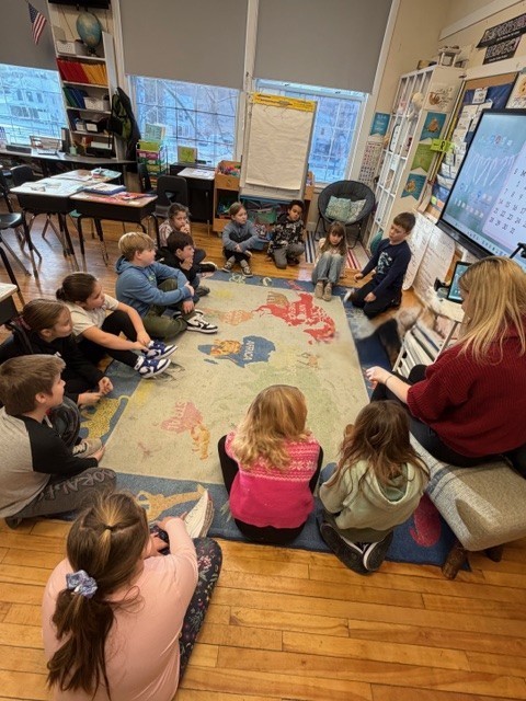 Elementary students sit in a circle on a classroom rug while a teacher leads a group discussion near a large interactive screen.