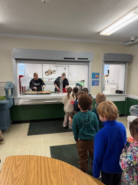 Students wait in line at the cafeteria while staff serve lunch behind the counter.