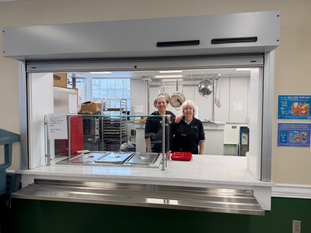 Two cafeteria staff members stand smiling behind the serving window in the kitchen.