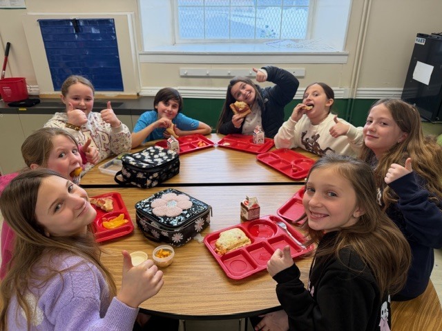Group of students sit around a table enjoying lunch and giving thumbs-up.