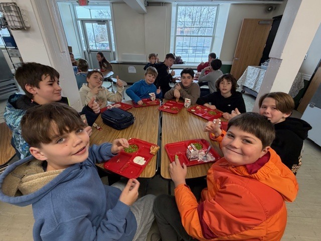 Students seated at cafeteria tables smile toward the camera while eating lunch.