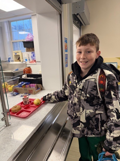 Elementary student smiles while holding a red lunch tray at the cafeteria counter.