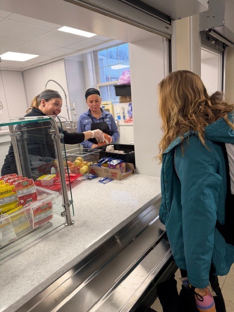 Students line up at the cafeteria serving window as staff hand out lunch trays.