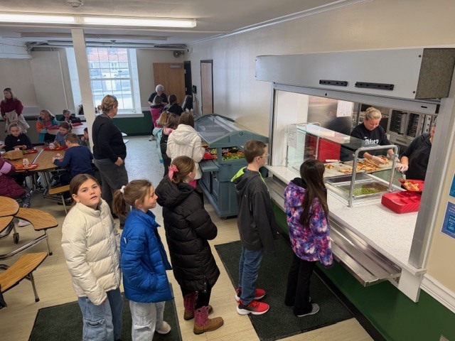 Wide view of cafeteria with students seated at tables eating lunch and chatting.