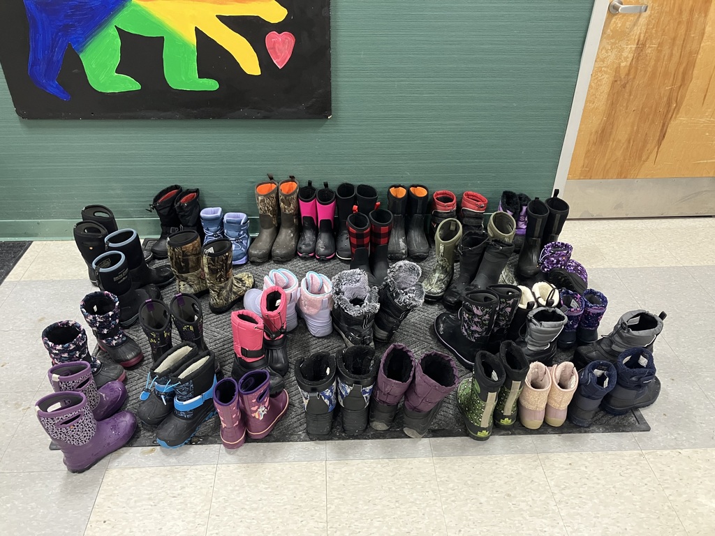 A row of children’s winter boots in many colors and styles is neatly lined up on a mat along a school hallway wall. The boots vary in size and pattern, showing they belong to multiple students.