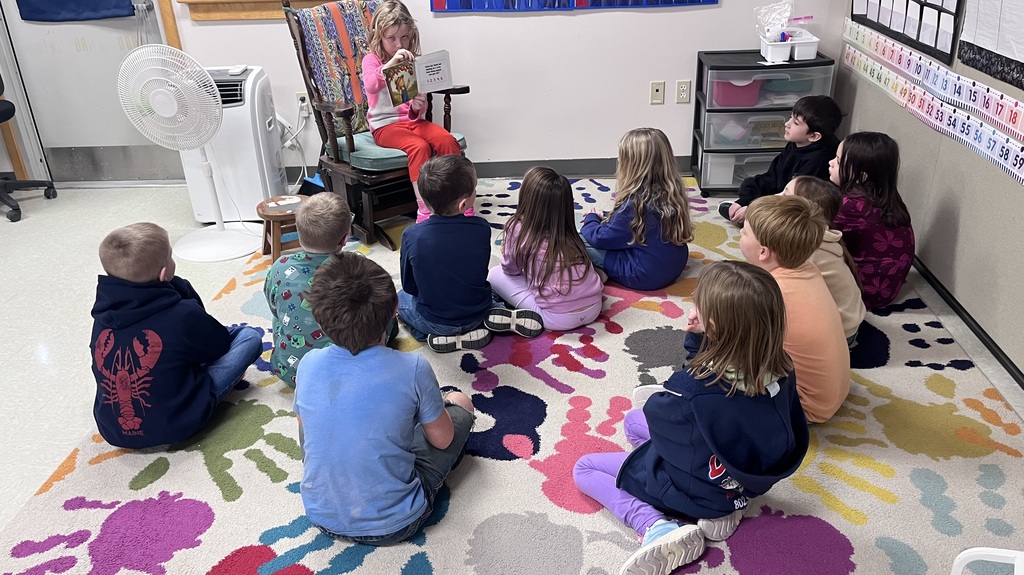 A young girl sits in a wooden rocking chair at the front of a classroom, reading a picture book aloud to a group of about nine children. The children are seated on a large, colorful rug decorated with multi-colored handprint patterns, all facing the reader. The classroom environment includes a white standing fan, an air conditioning unit, and educational materials like a number line and storage bins along the wall.