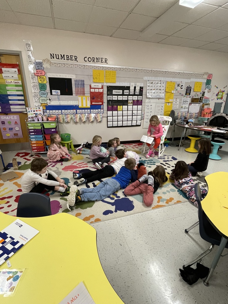 A wide shot of a classroom "Number Corner," where a young girl in a pink shirt and red pants sits in a white chair reading to her classmates. Nine other children are gathered on a large, colorful rug featuring multi-colored handprints; some sit upright while several others lie on their stomachs to listen. The wall behind them is filled with educational materials, including a January 2026 calendar, alphabet charts, and a "SHAPES" poster. Brightly colored classroom furniture, such as yellow tables and a rainbow-colored bin organizer, surrounds the group.