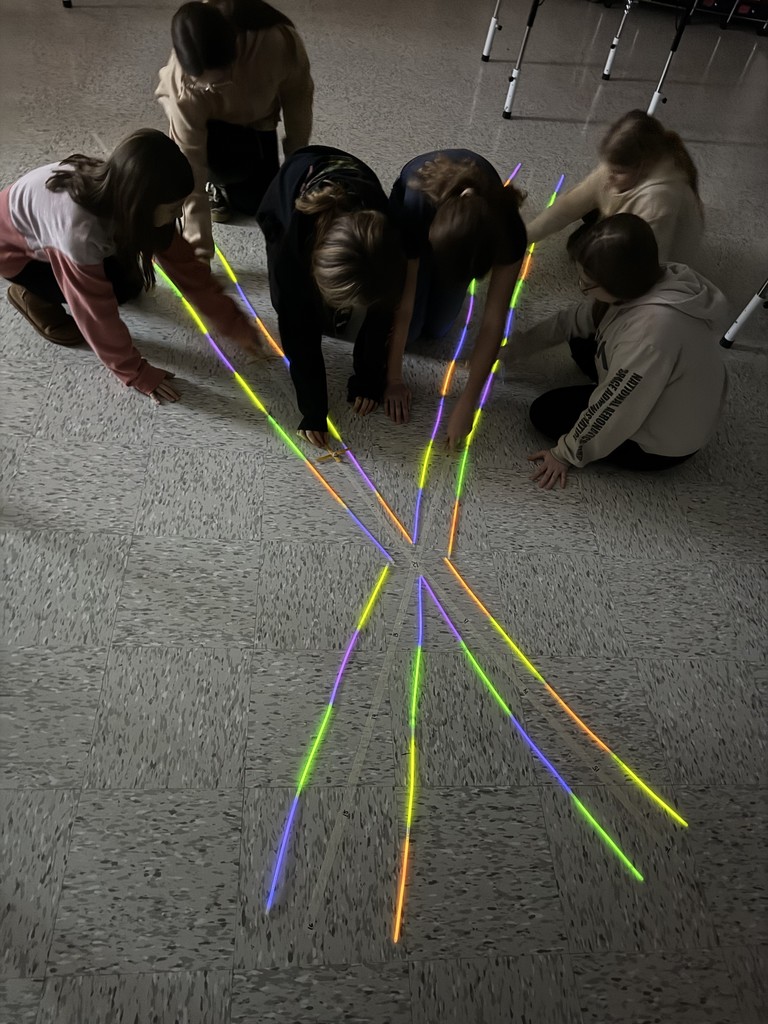 A group of elementary-aged students kneel on the classroom floor, working together to arrange colorful glow sticks into symmetrical, radiating lines that meet at a central point, creating a starburst pattern. The students lean in and collaborate as they carefully place the glow sticks, with classroom chairs visible in the background.