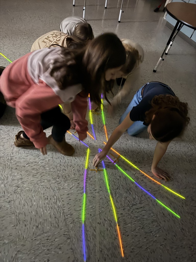 A group of elementary-aged students kneel on the classroom floor, working together to arrange colorful glow sticks into symmetrical, radiating lines that meet at a central point, creating a starburst pattern. The students lean in and collaborate as they carefully place the glow sticks, with classroom chairs visible in the background.
