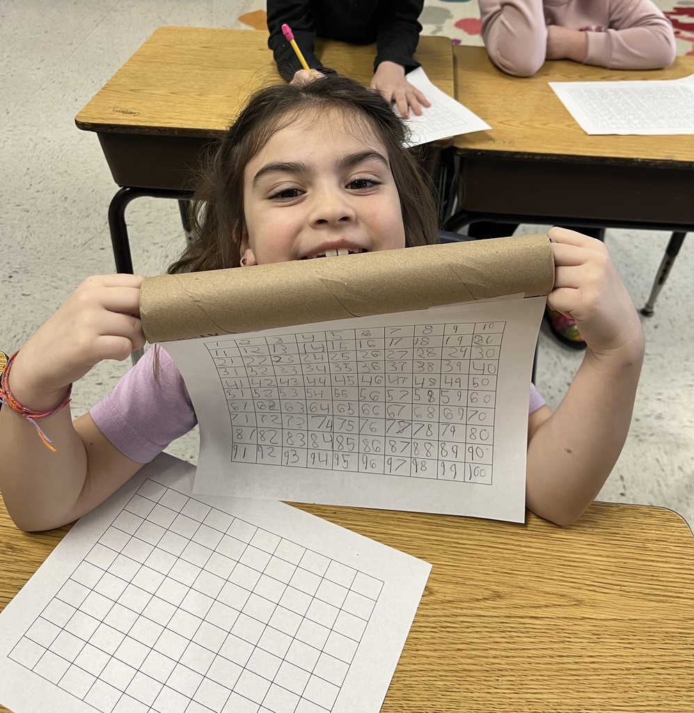 A young girl with dark hair and a purple shirt smiles broadly as she peeks over a cardboard tube. She is holding a sheet of paper with a handwritten 1 to 100 number grid taped to the tube to create a scroll. An empty grid sits on the desk in front of her. 