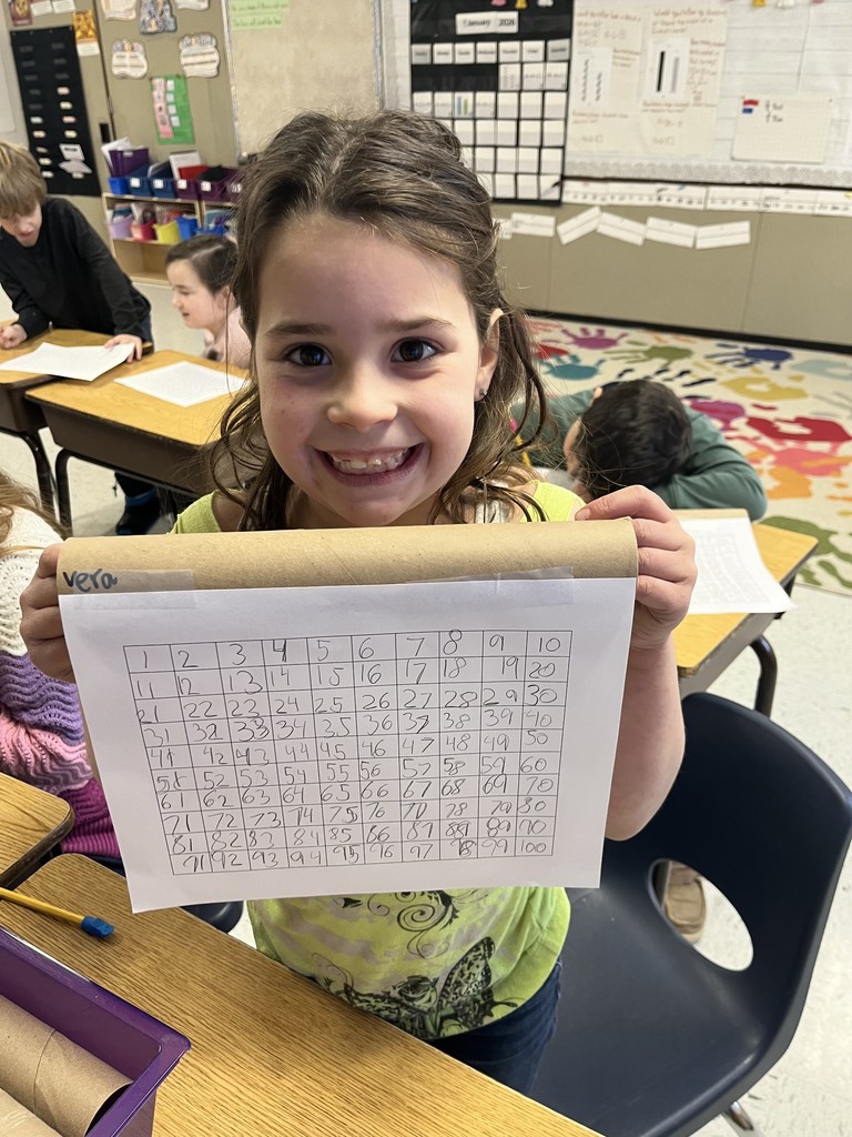 A young girl, wearing a bright green patterned shirt, smiles for the camera while holding up her number scroll. The scroll contains a completed 1 to 100 number grid handwritten in pencil. Other students and desks are visible in the background of the classroom.