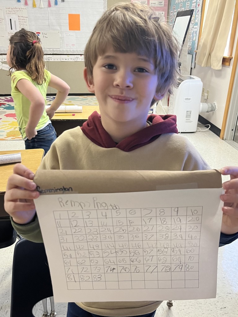 A young boy wearing a tan hoodie smiles while holding his number chart. His chart is filled out up to the number 83. The classroom background shows a colorful rug and another student.