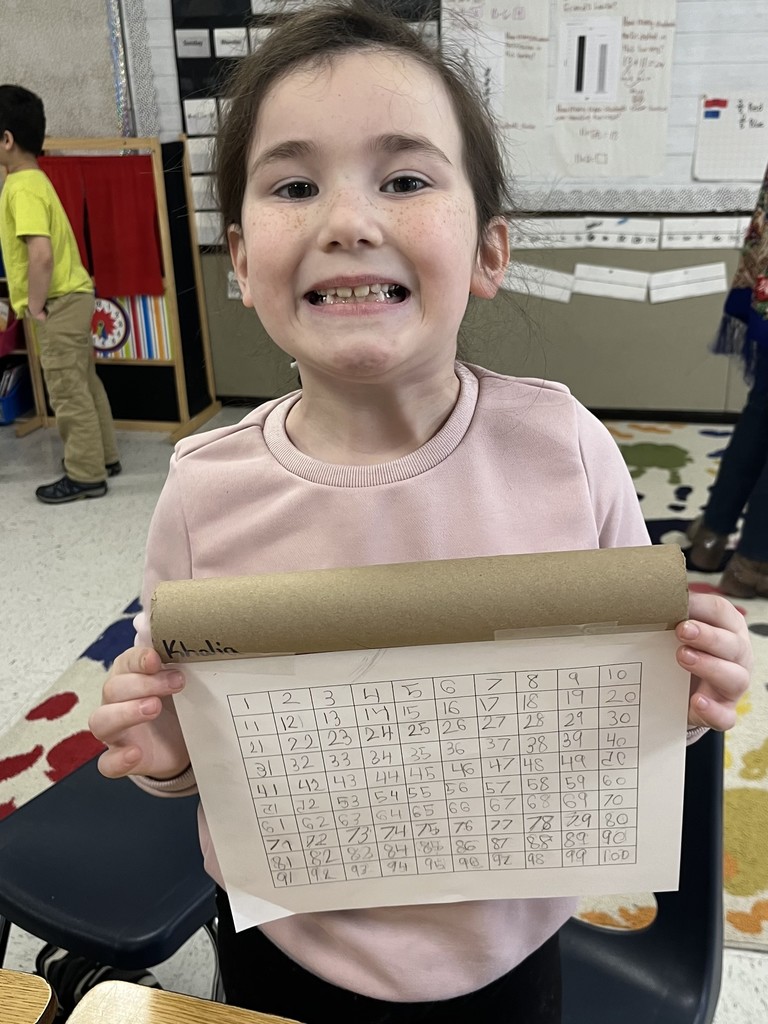 A smiling girl , wearing a light pink sweater, holds up her completed number grid. She is standing in a classroom with educational posters and a calendar visible behind her.