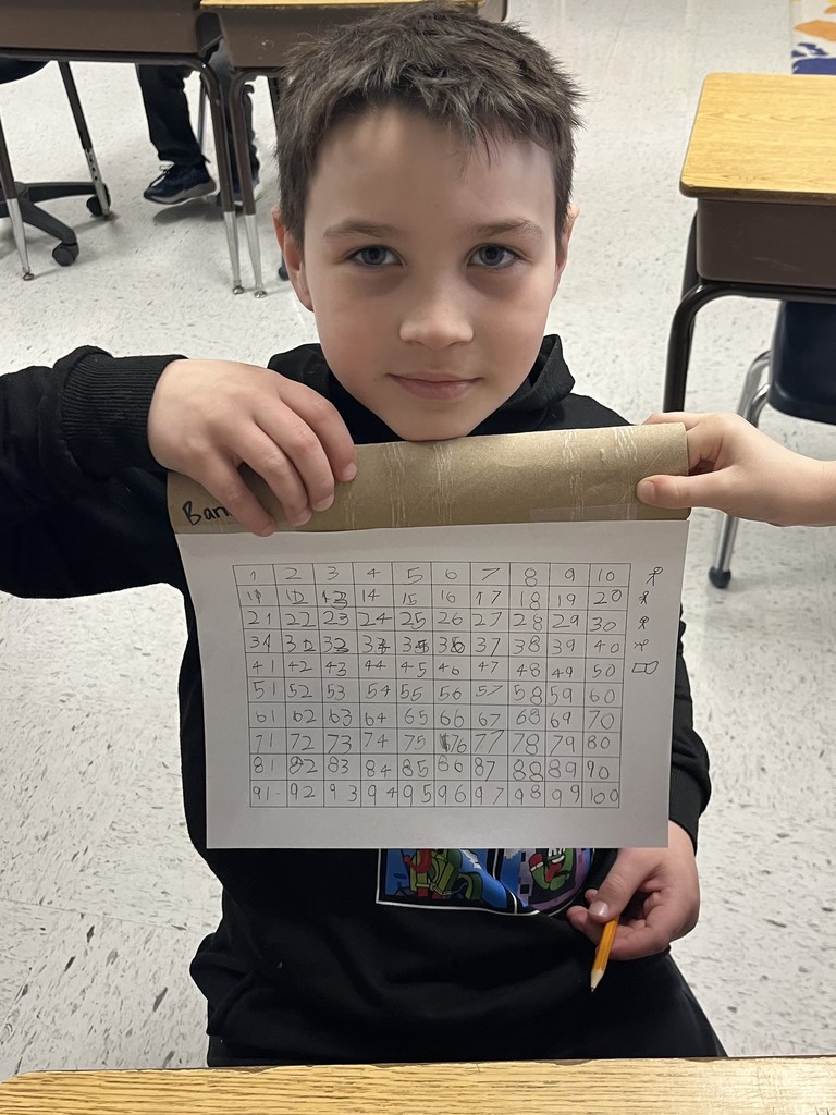 A boy in a black hoodie holds his completed hundreds chart. On the right margin of the paper, he has drawn small stick figures and a pair of glasses next to the rows of numbers.