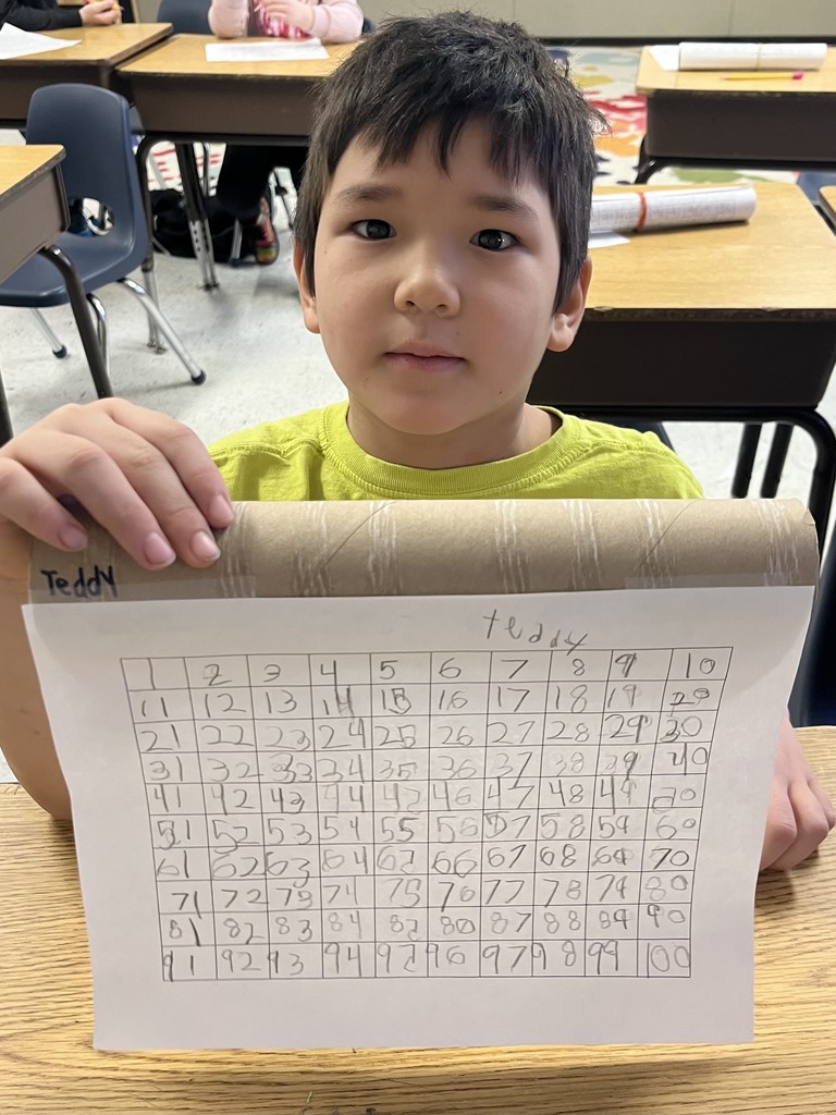 A boy named in a bright lime green shirt holds his completed hundreds chart while sitting at a school desk. The numbers are written clearly in pencil within the grid squares. IMG_3704 (Khalia): A smiling girl named Khalia, wearing a light pink sweater, holds up her completed number grid. She is standing in a classroom with educational posters and a calendar visible behind her.