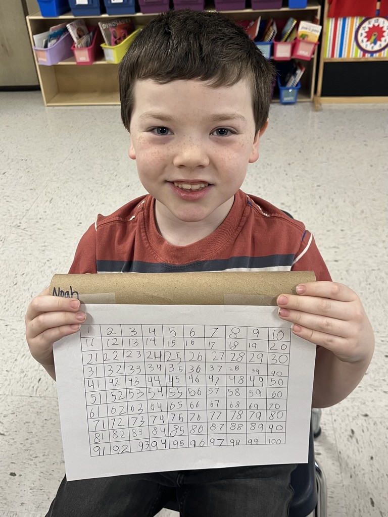 A boy with freckles , wearing a red and grey striped shirt, holds a fully completed 1-100 number grid. He is sitting in front of a shelf filled with colorful book bins. I