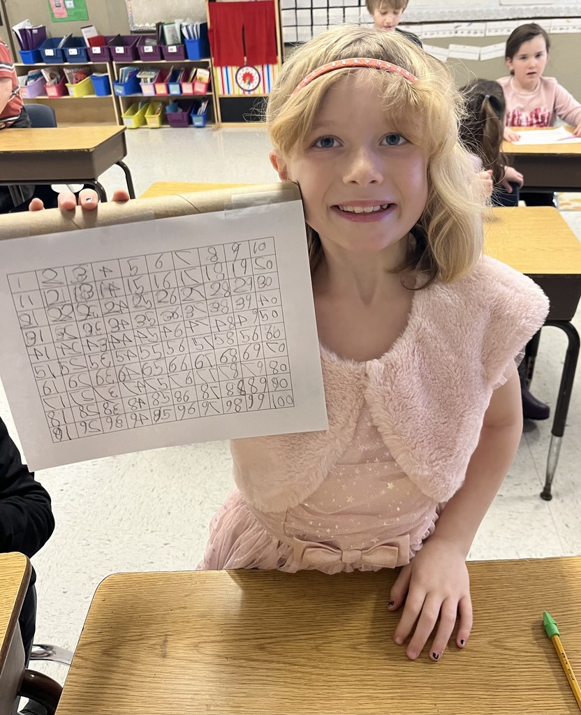 : A girl wearing a fancy pink dress with a sparkly headband holds up her hand-written number grid. She is standing by a wooden desk in the classroom.
