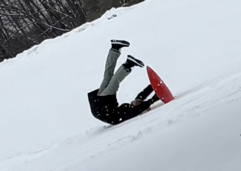 A student tumbles backward in the snow while sledding or snowboarding, with legs briefly in the air, capturing a lighthearted moment of winter play.