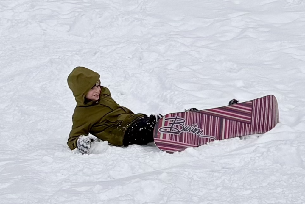 A student wearing a green winter jacket lies back in the snow with a snowboard attached, smiling after a playful fall on a snowy hillside.