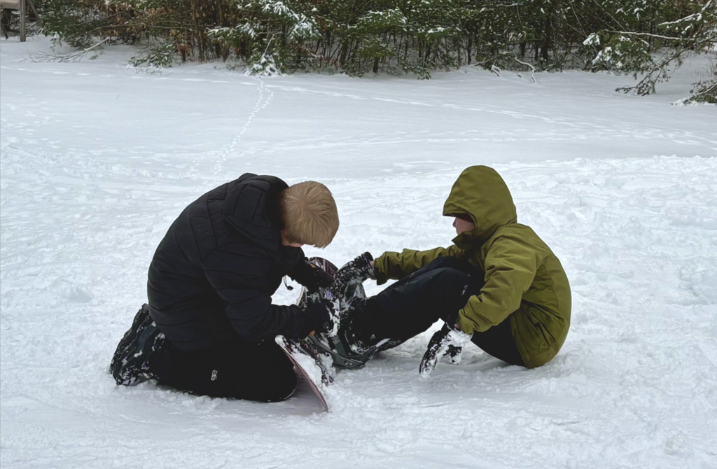 Two students sit in the snow facing each other, working together to fasten snowboard bindings, surrounded by a snow-covered field and trees.