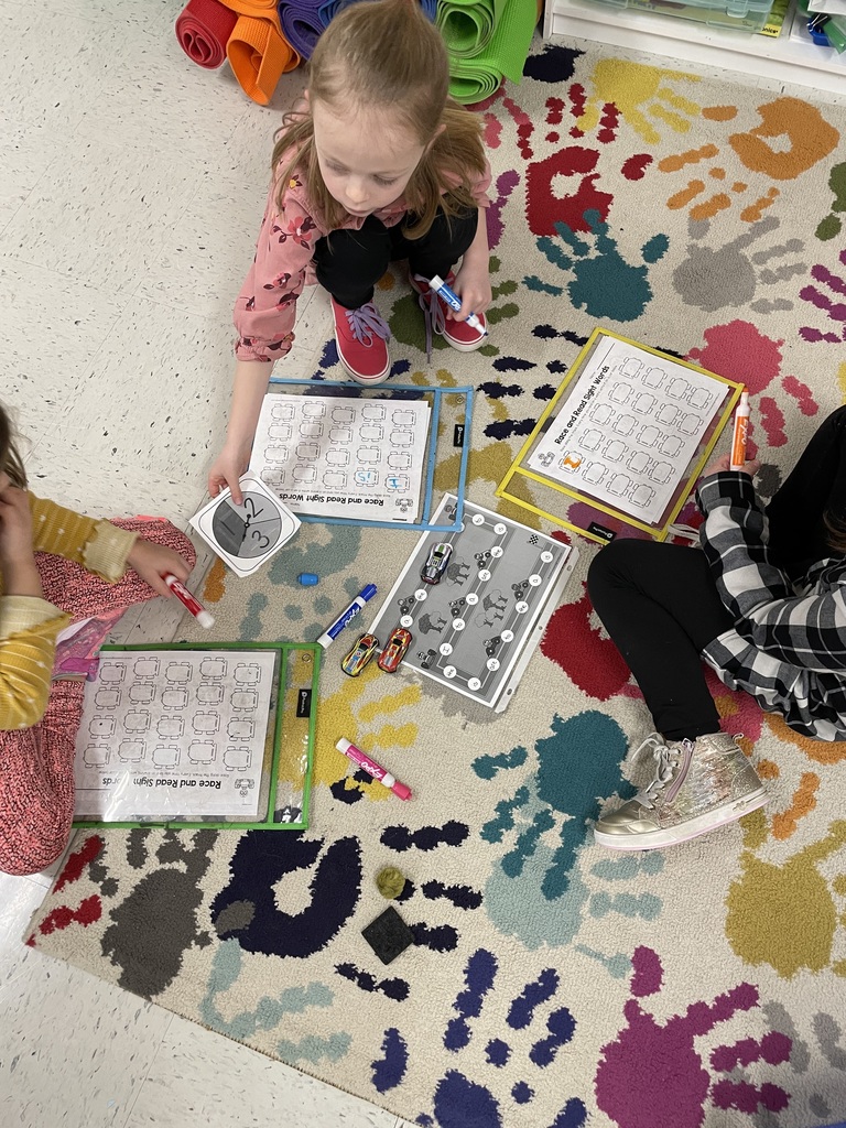 Three young children sit on a colorful rug decorated with handprints. They are using dry-erase markers and small toy cars to complete "Race and Read" sight word worksheets inside plastic sleeves. 