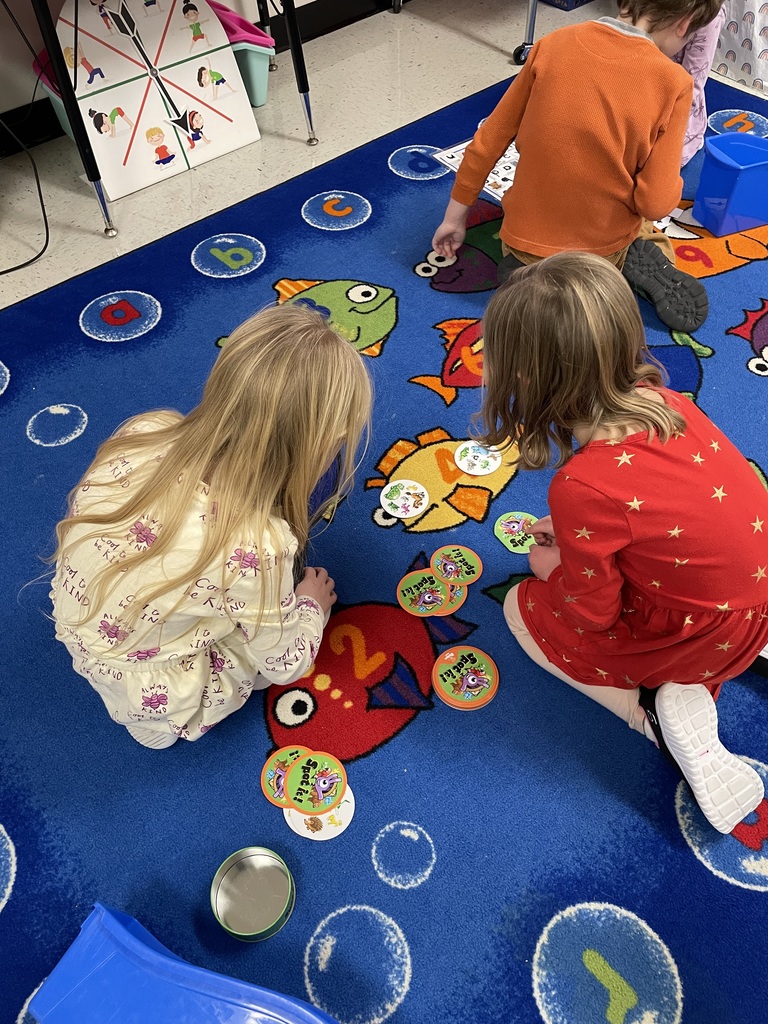  Two young girls sit on a blue classroom rug playing a game of "Spot It!" with circular cards. One girl is wearing a red dress with gold stars, and the other is in a cream-colored top.  