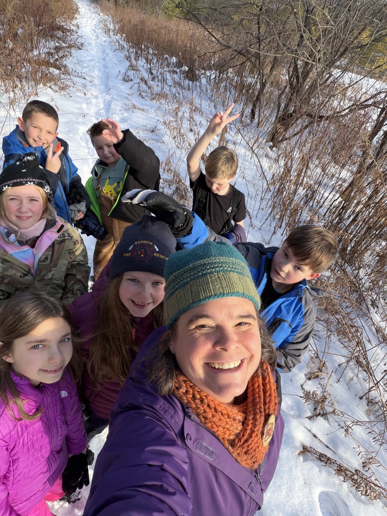 A group selfie of a smiling adult in a purple jacket and orange scarf with six children following behind her on a narrow, snowy trail.