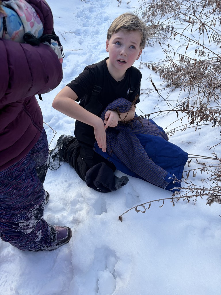 A boy in a black t-shirt and snow pants kneels on a snowy path, looking up and gesturing with his hands while his blue jacket lies on the snow beside him.