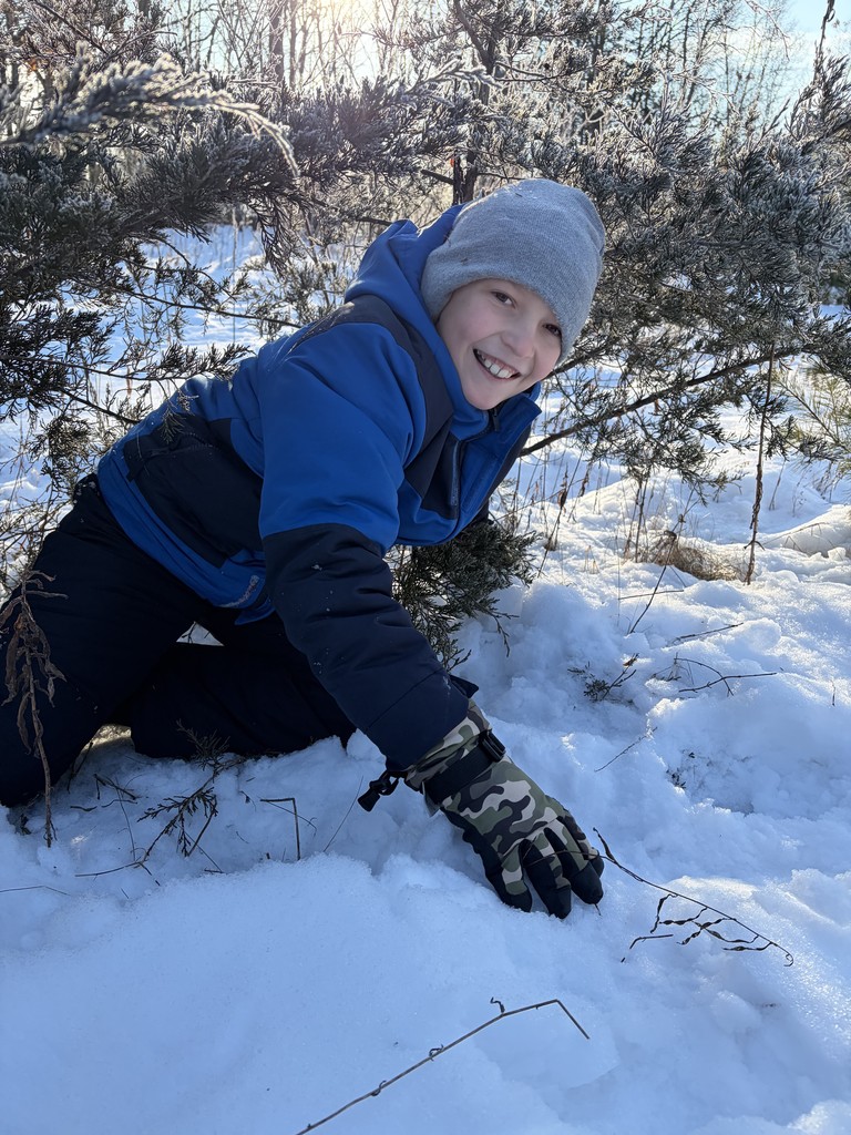 A boy wearing a blue and black jacket and camouflage gloves smiles brightly while kneeling in the snow beneath frost-covered evergreen branches.