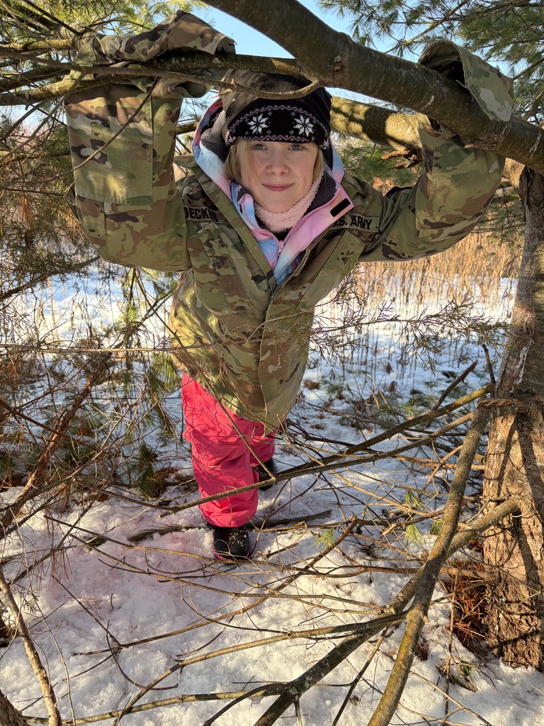 A smiling girl wearing an oversized camouflage U.S. Army jacket and pink snow pants, standing behind pine tree branches.