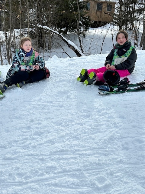 Two students sit on the snow next to snowboards, smiling while taking a break on a gentle slope.
