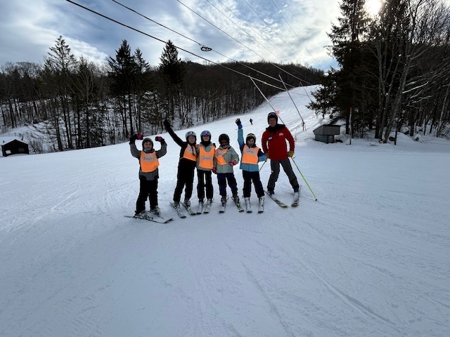 A group of young skiers wearing orange vests pose with an instructor on a wide ski trail, raising their arms in celebration.