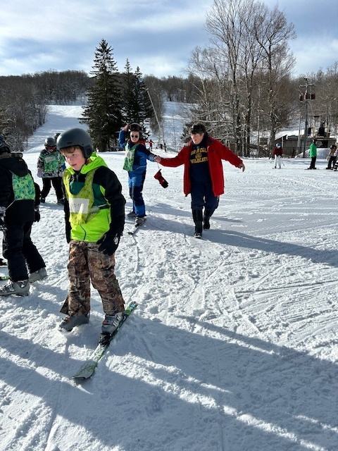 Elementary students wearing helmets and skis glide slowly across a snowy beginner area while instructors supervise nearby.