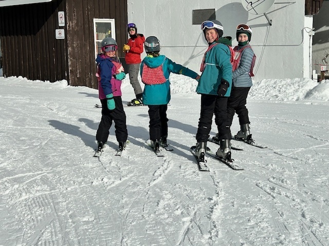 Four students in winter gear stand on skis near a building, looking back toward the camera during a break in their lesson.