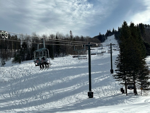 A chairlift moves uphill over a snowy ski trail surrounded by trees under a partly cloudy sky.