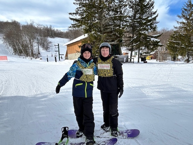 Two elementary students stand side by side on snowboards, smiling at the camera on a groomed ski area.