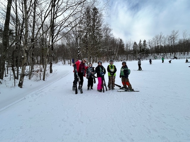 A group of elementary students and an instructor stand together on skis at the edge of a ski trail, preparing to continue their lesson.