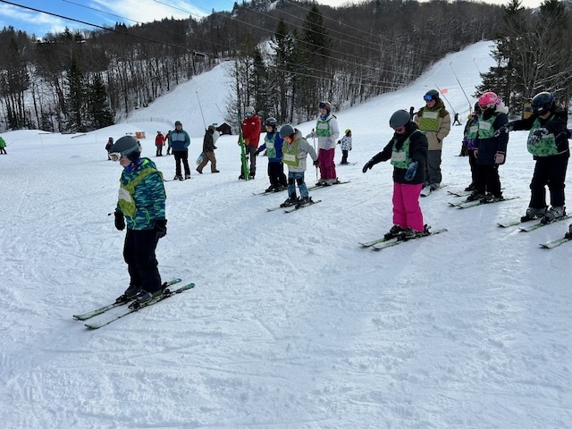 A line of young skiers practices standing and moving forward together on a snow-covered slope during a ski lesson.