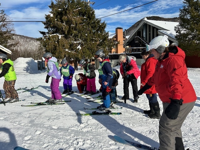 A group of elementary students in helmets and skis line up on a snowy slope while instructors in red jackets help them prepare to start skiing.
