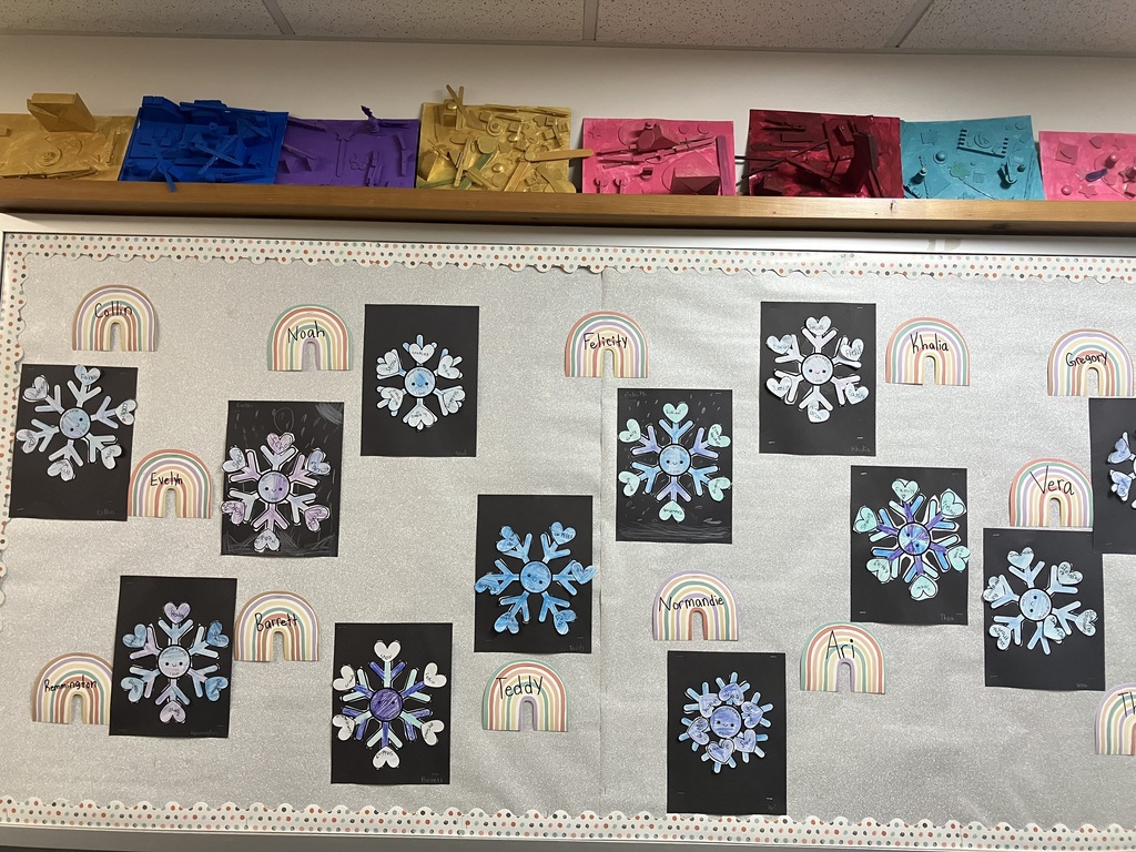  Classroom Bulletin Board  A wide shot of a classroom bulletin board covered in silver glitter paper. It displays several student art projects: paper snowflakes on black backgrounds and rainbow-shaped name tags. Above the bulletin board, a wooden shelf holds a row of monochromatic 3D assemblages made from various objects, each painted a single bright color like gold, blue, purple, red, and teal. 
