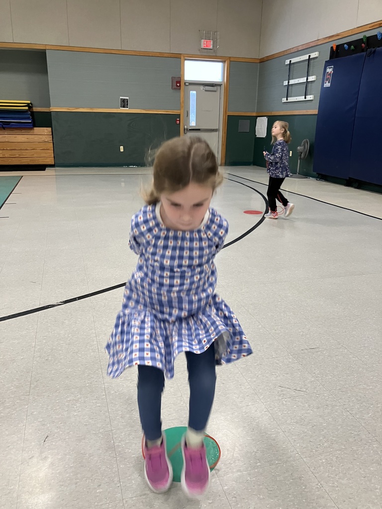 A young girl in a blue and white checkered dress jumps over a green circular floor marker in a gym.