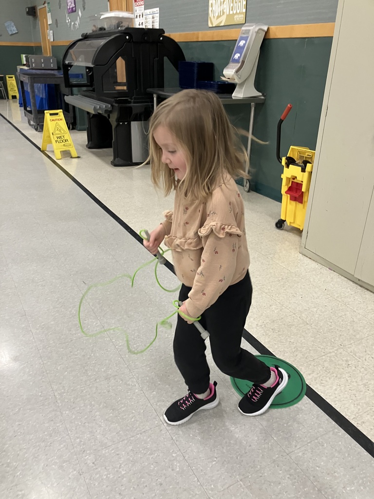 A young girl in a tan ruffled sweatshirt and black leggings walks across the gym floor while holding a green jump rope.