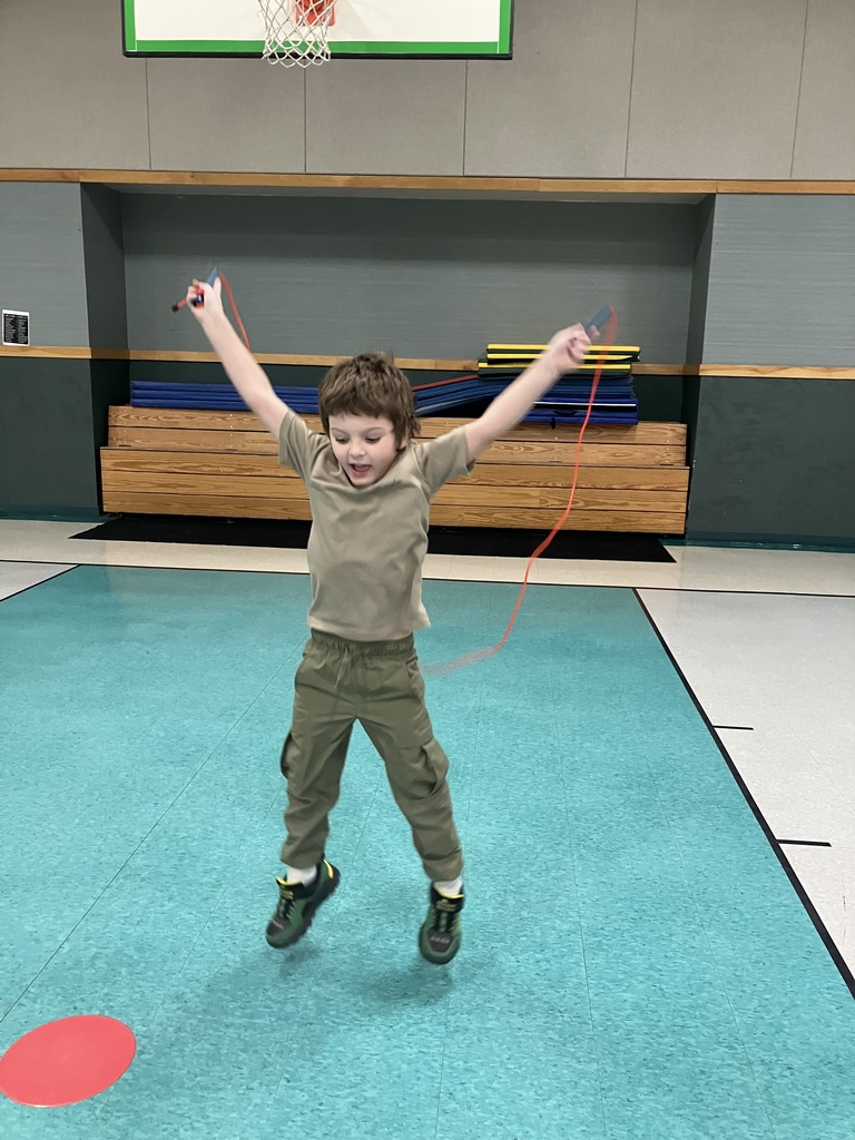 A young boy in a tan t-shirt and olive cargo pants jumps high into the air with his arms raised while using a red jump rope.