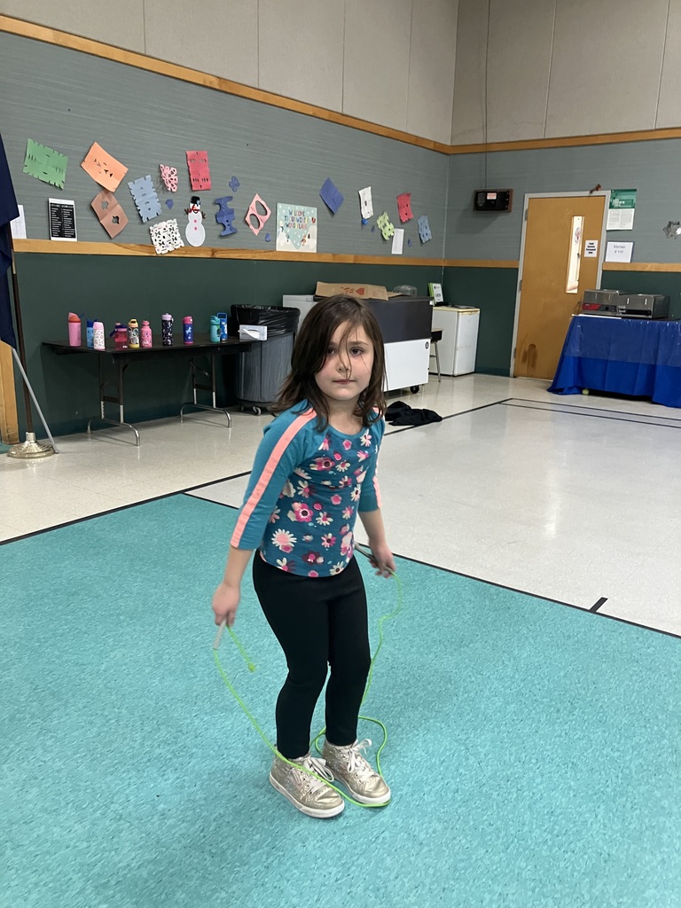 A young girl with long brown hair, wearing a teal floral shirt and black leggings, stands ready with a green jump rope on a blue section of the gym floor.
