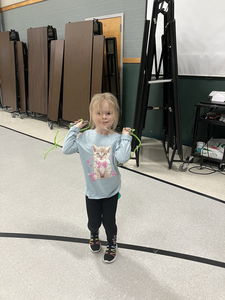 A smiling young girl in a light blue shirt featuring a kitten graphic holds a green jump rope behind her shoulders.