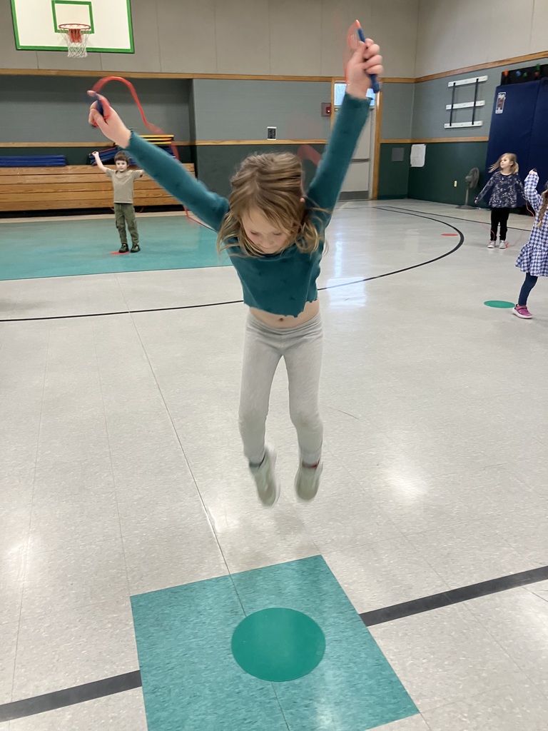 A young girl in a teal long-sleeve shirt and grey leggings is captured mid-air while jumping with a red jump rope in a gymnasium